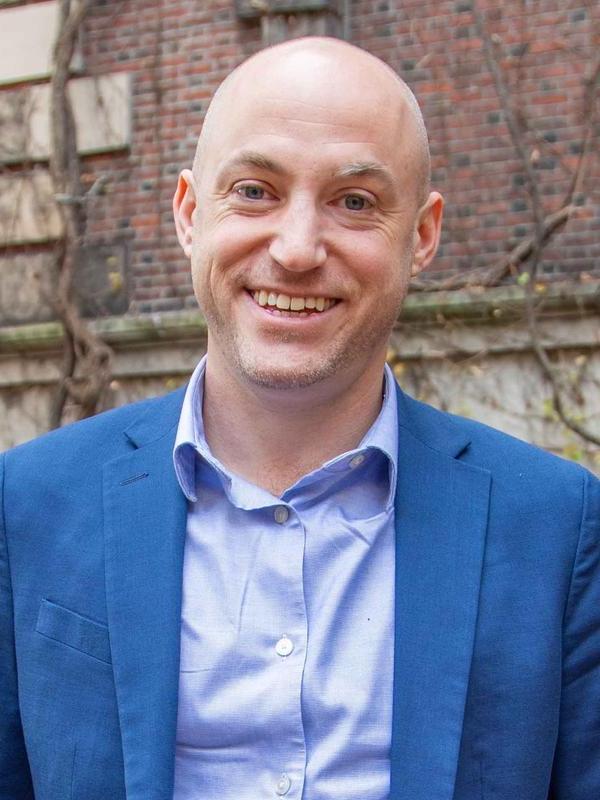 a - j Aronstein穿着一件有领的纽扣衬衫(淡紫色)和一件蓝色运动夹克，在一座棕色建筑前微笑着